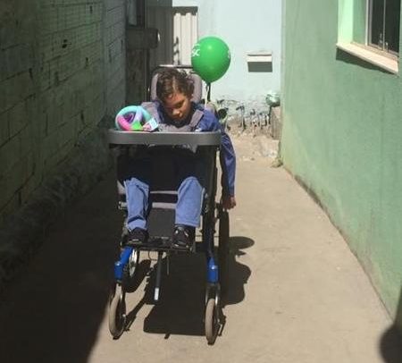 Garoto de cerca de 10 anos está na cadeira de rodas em um corredor. Ele é castanho claro, tem cabelos encacheados, veste roupas azuis, e tem uma bexiga verde presa na cadeira.