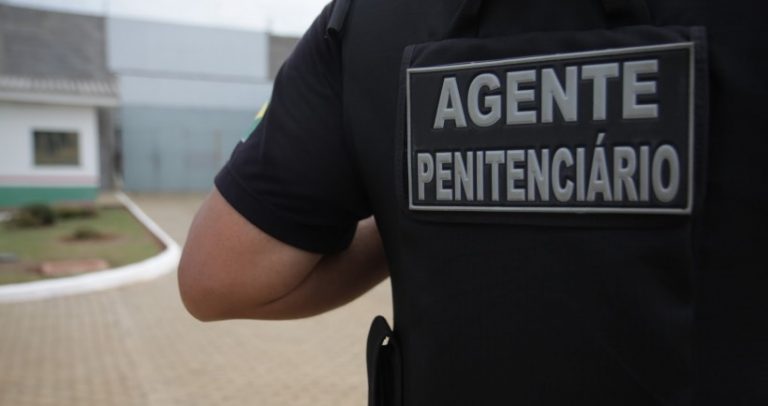 Descrição da imagem: agente penitenciário aparece de costas e na sua farda preta está escrito em cinza Agente Penitenciário