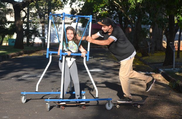 Descrição da imagem: Ricardo Porva, de boné, calça bege e camiseta preta. Sobre um skate ele empurra o equipamento onde está sua filha de 13 anos, amarrada por elásticos e segurando nas bordas da "gaiola" azul e branca. Ela também desliza sobre um skate. Ela tem a pele clara, cabelos compridos e sorri.