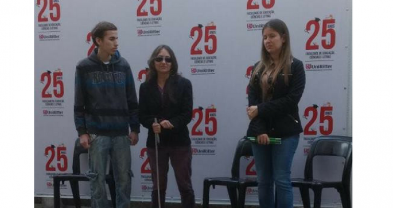 Na foto vertical e colorida, da esquerda para a direita estão Gabriel Pessoa Ribeiro, Franciele Brandão e Mariana Baierle. Eles estão em pé. Franciele usa óculos escuros e segura sua bengala branca aberta. Mariana segura sua bengala verde dobrada. Atrás deles, há três cadeiras plásticas pretas. Atrás das cadeiras, uma parede branca apresenta várias vezes a mesma logomarca com o seguinte conteúdo textual: "25 anos Faculdade de Educação, Ciências e Letras Uniritter". Na logomarca, o número 25 está escrito em vermelho e todas as palavras, em preto.