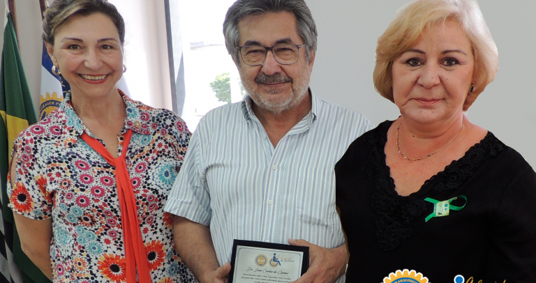Descrição da imagem: Dr. Kal segura a placa em sua homenagem, ladeado por Marisa e Rose, ambas loiras e sorridentes