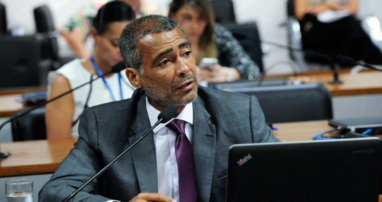 Na imagem o senador Romário está no plenário falando ao microfone. Ele é mulato, usa terno escuro e camisa branca