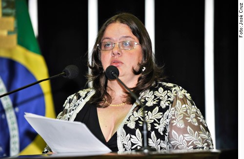 Descrição da imagem: Adriana Dias fala ao microfone segurando um papel. Ela tem a pele clara, cabelos castanhos na altura dos ombros e usa óculos