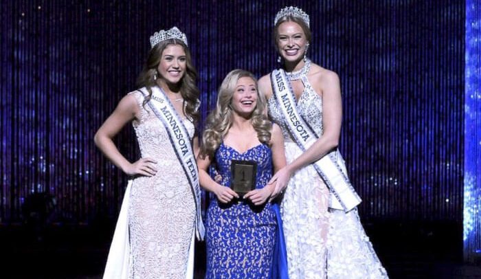 Descrição da imagem: Mikayla está de vestido longo azul, de rendas, entre duas misses vestidas de branco. Ela sorri carregando um troféu. Todas são loiras