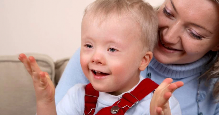 Descrição da imagem: Uma mãe segura no colo seu filho com síndrome de Down. Ambos são loiros. Ele tem cerca de 2 anos e usa blusa branca e macacão vermelho por cima. Eles sorriem.