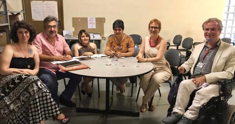 Descrição da imagem: Seis integrantes da Câmara para Inclusão sentados numa mesa redonda. Dr. Kal é o segundo à esquerda. Ele veste camisa cor de rosa, usa barba grisalha e óculos. Todos olham para a câmera e sorriem.