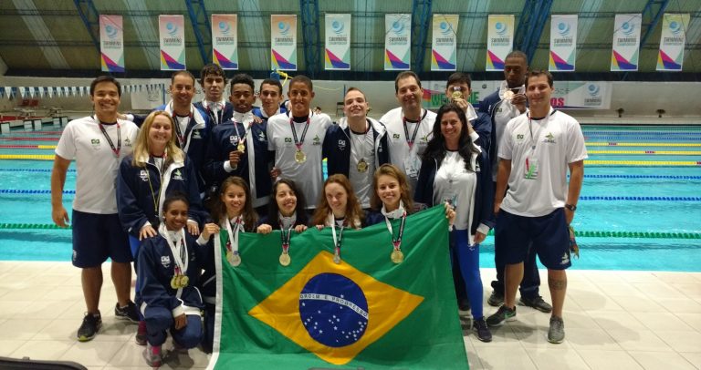 Descrição da imagens: 13 atletas com uniforme da seleçãobrasileira estão em frente a uma piscina onde participaram do campeonato mundial no México. Na frente deles, a bandeira nacional
