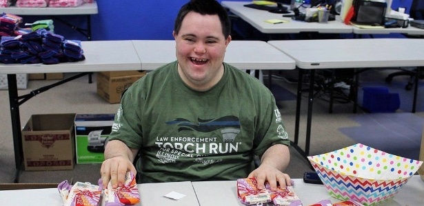 Descrição da imagem: John, de 23 anos, está numa mesa, sorrindo e segurando alguns pacotes de meia. Ele usa camiseta verde escura, tem a pele clara, cabelos castanhos e sorri.