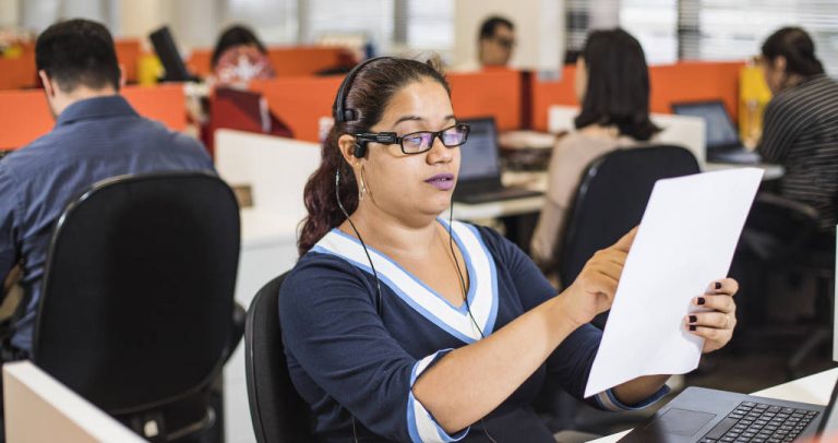 Descrição da imagem: Mulher trabalha com fone de ouvido e óculos com câmera para leitura de textos. Ela tem a pele clara, os cabelos castanhos e usa blusa azul marinho e branca