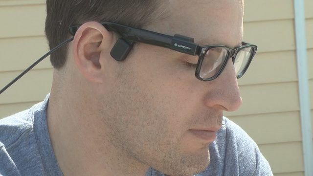 Descrição da imagem: Homem usa óculos com o dispositivo orcam my eye na aste do óculos. Ele tem a pele clara e os cabelos castanhos e está de perfil.