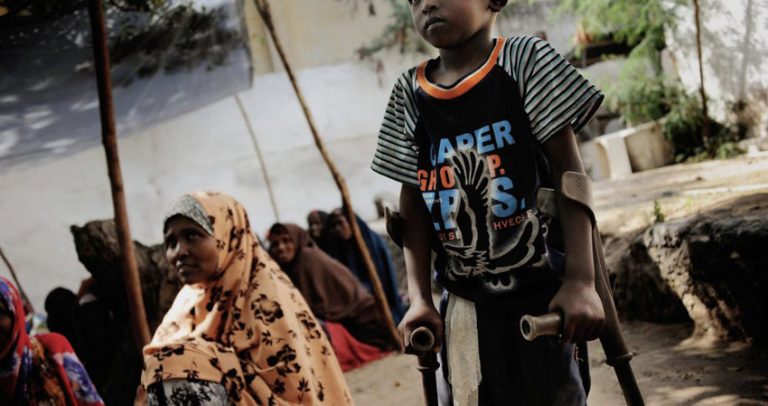 Foto de um menino de pele negra andando com o apoio de muletas. Atrás dele aparecem mulheres sentadas no chão, sorrindo e usando roupas de tecidos estampados que cobrem a cabeça.
