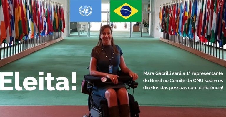 Foto de Mara Gabrilli na ONU em Nova York. Ela está em sorridente no centro do salão onde ficam as bandeiras de todos os países, uma ao lado da outra. Em destaque, ao fundo, estão as bandeiras do Brasil e a bandeira da ONU.