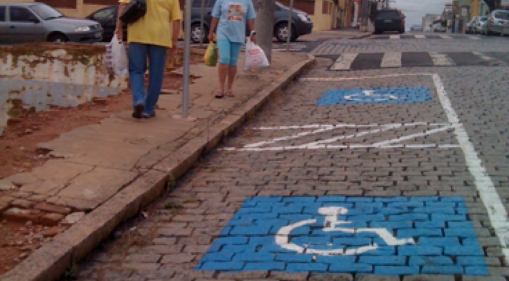 A mobilidade depende de espaços acessíveis. Foto de calçada e rua de paralelepípedo com demarcação de vaga, com o símbolo da pessoa com deficiência. Porém a calçada está toda danificada, não há guia rebaixada nem área de transferência na vaga.