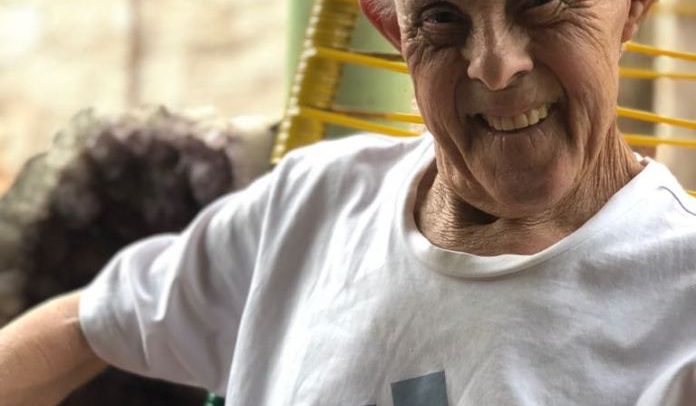 Foto de João sorrindo, sentando em uma cadeira. Ele tem cabelos brancos.