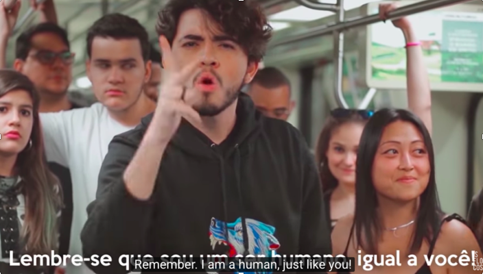 descrição da imagem: foto de um moço fazendo sinais em Libras, com expressão séria. Ele está dentro de um ónibus e atrás dele tem várias pessoas jovens.