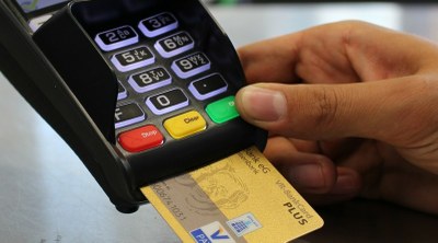 Fotografia mostra parte de uma mão segurando uma máquina de pagamento, com destaque para o teclado e um cartão inserido.