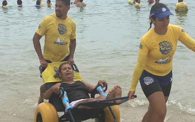 Foto de dois voluntário vestidos com camisas amarelas e shorts pretos, saindo do mar e puxando uma mulher em uma cadeira de rodas especial, com o assento de material permeável e de rodas largas. Eles parecem estar conversando.