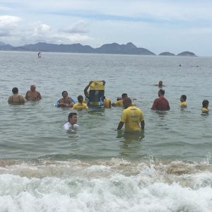 foto de várias pessoas com deficiência e voluntários vestidos com camisas amarelas. Todos estão perto da praia, com o mar à altura do peito.
