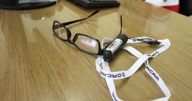 Fotografia do óculos Orcam, que vem acoplado com um equipamento scanner portátil. Aparentemente, ele é similar a um óculos convencional.