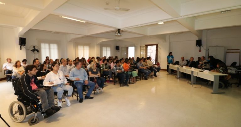 Fotografia mostra audiência do Seminário, onde estão dezenas de pessoas. Em destaque, do lado esquerdo, um participante em cadeira de rodas tendo ao fundo, do lado direito, a mesa de trabalhos