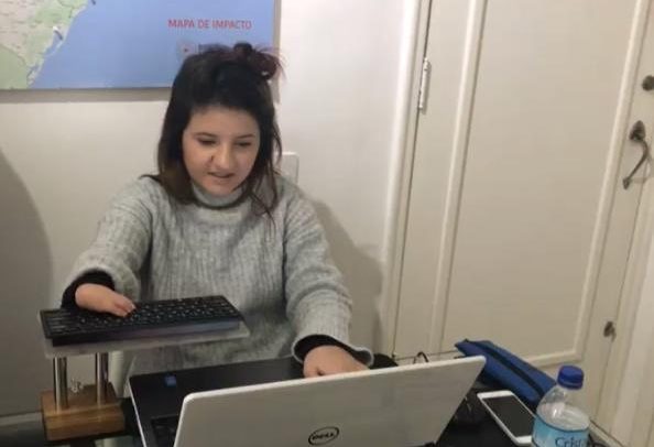 Duda está sentada diante do notebook, e tem um teclado mais próximo do seu corpo para facilitar o alcance. Ela tem um encurtamento de membro no braço.