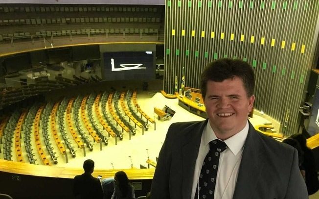 Felipe Rigone aparece sorrindo (lado direito da fotografia), tendo como cenário, ao fundo, o plenário onde acontecem os debates, votações e homenagens da Assembleia Legislativa, em Brasília