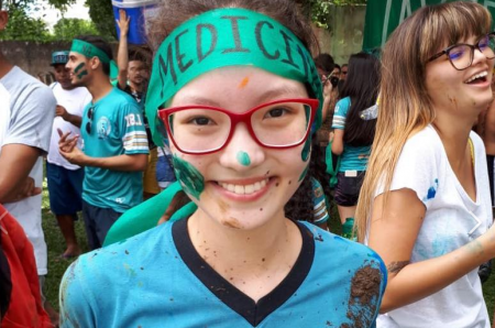 Fotografia mostra Natãmy, com colegas ao fundo. Com sorriso nos lábios ela usa na testa a faixa de caloura de medicina. O resto está parcialmente pintado com tinta colorida na testa, nariz, bochechas e queixo, com fragmentos de lama, devido ao trote de recepção na universidade