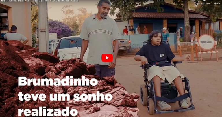O jovem Lázaro está acionando sozinho sua cadeira de rodas. Seu pai caminha ao seu lado, sorrindo. A rua por onde andam está repleta de terra.
