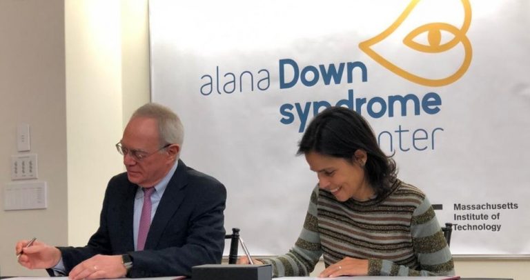 Ana Lúcia Villela está sentada ao lado do presidente do MIT, Rafael Reif. Ambos assinam o documento de doação. Ao fundo, a logomarca do alana Down Syndrome Center