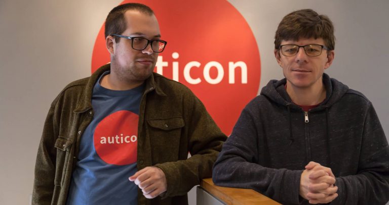 Fotografia mostra dois homens, um do lado do outro, com os braços apoiados numa divisória. Ao fundo a logomarca da empresa Auticon