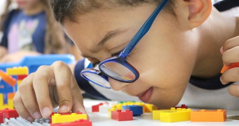 Fotografia mostra criança com baixa visão, usando óculos na cor azul, brindando com LEGO