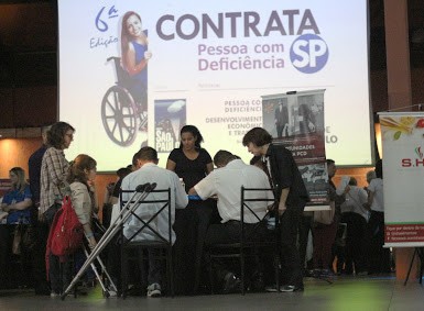 Pessoas com Deficiência são atendidos durante ä Edição do Contrata SP. Na fotografia três candidatos aparecem sentados, de costas, em frente ao painel com a logomarca oficial do evento. Três orientadores aparecem de pé, apoiando-os