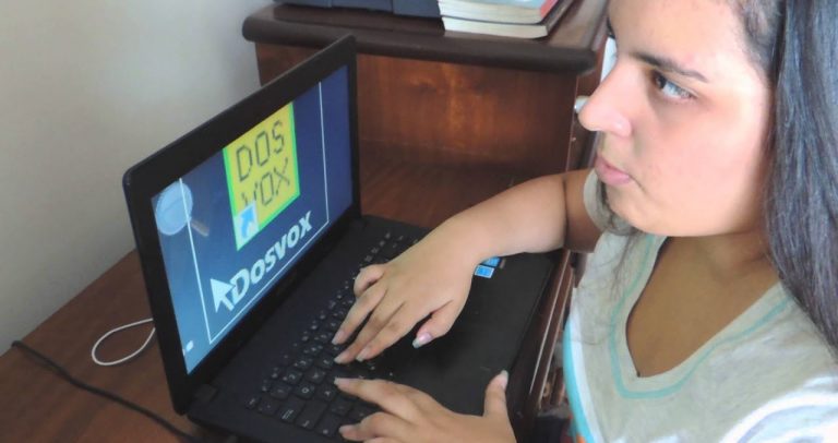 fotografia mostra jovem cega usando o computador. Ela está digitando e a tela mostra um programa aberto