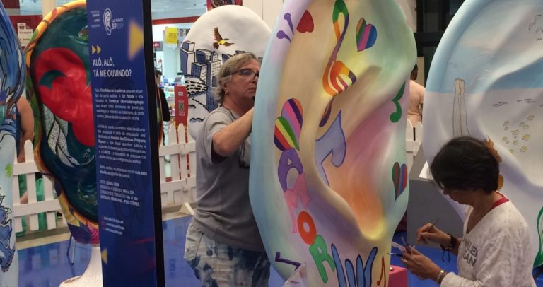 fotografia mostra detalhe da exposição Ear Parece. No lado direito, no meio das esculturas em forma de orelhas, placa explicativa na cor azul resume o evento, sob o título Alô Alô Tá me Ouvindo? E do lado direito duas artistas plásticas pintam com tintas coloridas a base branca, formando uma orelha com referências musicais