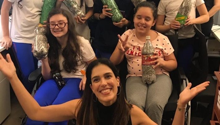 Fotografia mostra Carolina sorrindo e com as mãos para cima, em frente a um grupo de alunos com e sem deficiência. Os estudantes mostram projeto realizado com garrafas PET