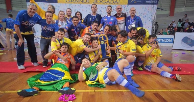 equipe técnica e atletas posam para fotografia festiva em frente a painel onde se lê Diversidade, uma grande jogada. Todos sorriem e fazem poses informais com as bandeiras e medalhas