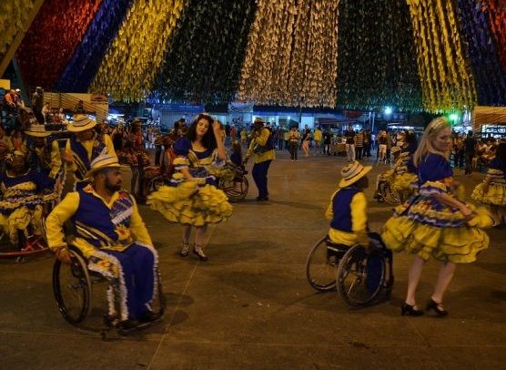 Fotografia mostra a bailarinos dançando a quadrilha. Em destaque, dois homens em suas cadeiras de roda ao lado de suas parceiras. Eles vestem trajes típicos das festas juninas e o show acontece em ambiente de decoração típica, com bandeirolas e luzes enfeitando o recinto de festa