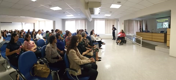 foto ilustrativa da reunião da Câmara Paulista mostra o auditório cheio de pessoas que ouvem uma das palestrantes, Sandra. Ela está sentada em sua cadeira de rodas, em frente ao público e tem ao seu lado esquerdo o intérprete de Libras. Ao fundo, detalhe do palco onde aparece uma mesa de trabalho e um telão