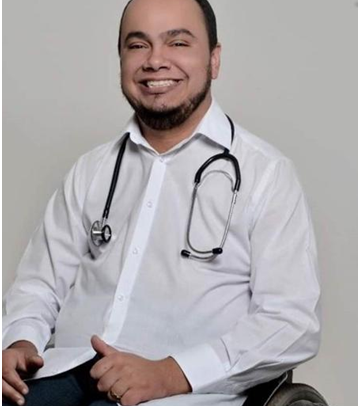 Fotografia do Dr sorrindo. Usando camisa branca e com o estetoscópio em torno do peito, ele está sentado em sua cadeira de rodas