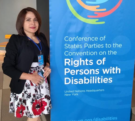 Vestindo vestido branco com estampa de flores vermelhas com detalhes e blazer na cor preta, Marinalva posa ao lado do banner do evento, que tem fundo azul e dizeres em outro tom de azul e branco: Conference of States Parties to the Convention on the Rights of Persons with Disabilities, United Nations Headquarters New York, WWW.un.org/disabilities #COSP #SDGs #GlobalGoals. No alto, a logomarca colorica simula um abraços em forma de círculos e embaixo a logomarca da ONu, na cor branca, e dos Objetivos do Desenvolvimento Sustentável, colorido
