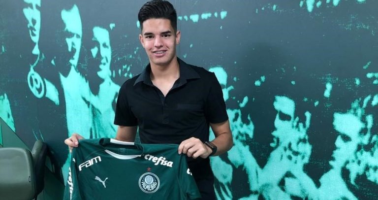 Posando em frente a um painel com fotografia de ex-jogadores, o Gustavo Peralta posa para fotografia segurando nas mãos a camisa do Palmeiras