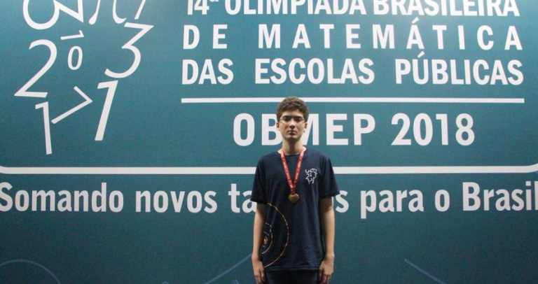 Gabriel posa com a medalha no peito em frente do painel oficial. Em fundo azul, onde aparece a logomarca do evento, está escrito: 14ª. Olimpíada Brasileira de Matemática das Escolas Públicas. OBMEP 2018 Somando novos talentos para o Brasil