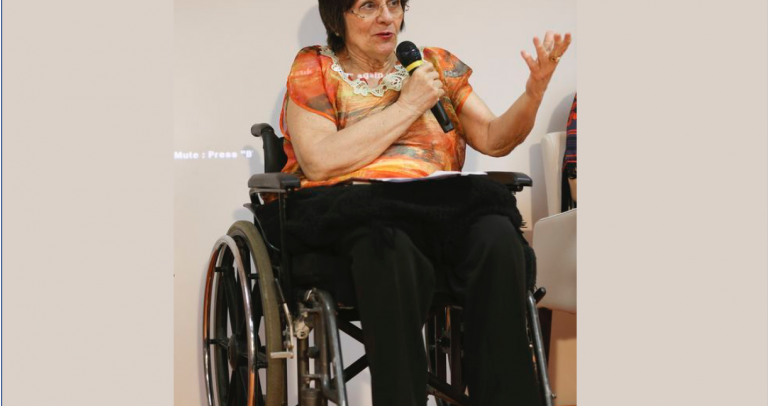 Maria da Penha fala ao microfone. Ela usa cadeira de rodas.