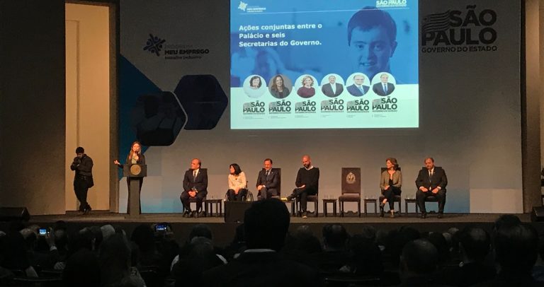 Palco onde estão o governador do Estado de São Paulo, João Doria e o prefeito da cidade de São Paulo, Bruno Covas, com 5 secretários estaduais. Entre os secretários está Célia Leão, da Secretaria dos Direitos das Pessoas com Deficiência. Ela usa cadeira de rodas.