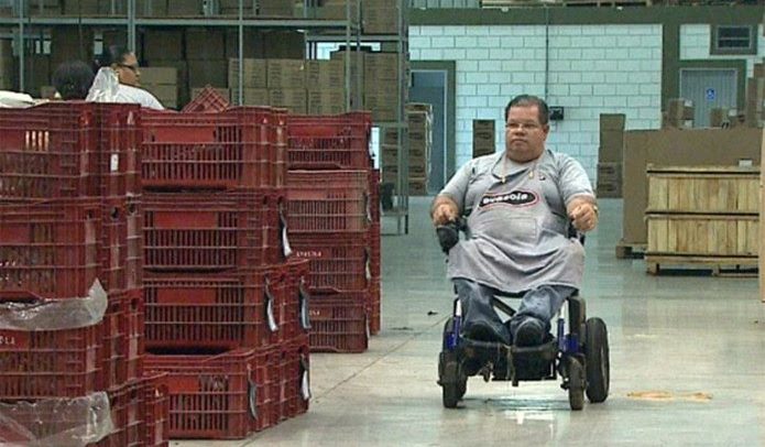 foto de um homem na cadeira de rodas, com uniforme de empresa, dentro de um galpão com muitas caixas empilhadas.