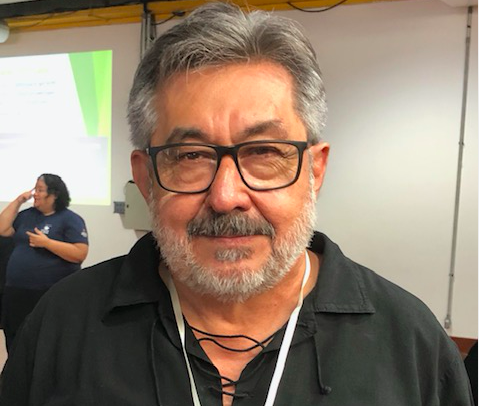 Foto de frente do Dr. José Carlos do Carmo, kal., da cintura para cima. Ele é um homem de cabelos grisalhos, usa óculos, barba curta, e está com uma camisa preta.