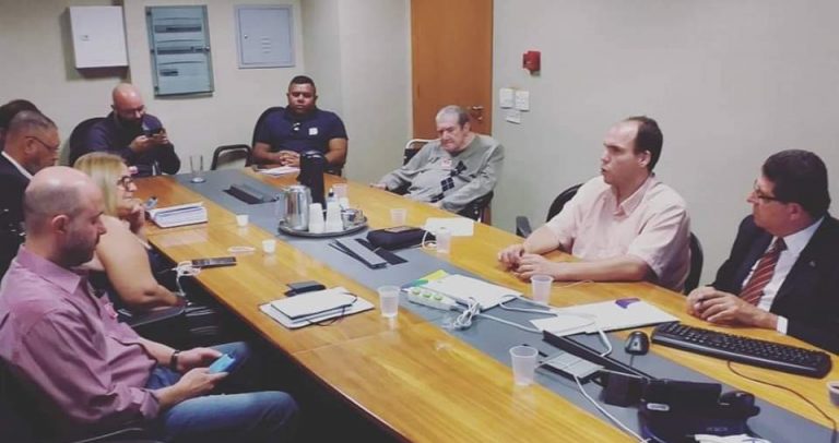 Foto da Comissão Organizadora da V Conferência reunida, feita a partir do canto esquerdo da sala. Os membros estão sentados em torno de mesa oval de madeira clara com faixa preta central em toda a sua extensão.