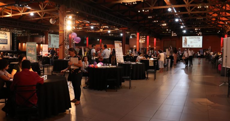 Foto das mesas das empresas e mais de 20 pessoas espalhadas no salão da Expo Barra Funda. Funcionários atendem os participantes.