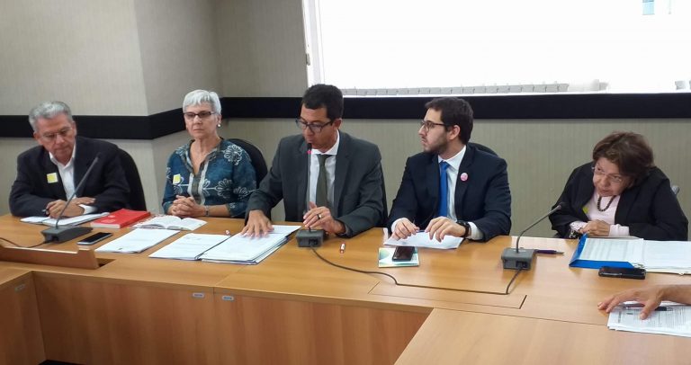 Foto da mesa diretora do Plenário do Conade durante o debate sobre o Probad. Da esquerda para a direita, sentados atrás da bancada de madeira clara, Dr. Heleno Rodrigues Corrêa Filho, de paletó escuro e camisa clara sem gravata; Dra. Maria Aparecida Gurgel, de blusa verde com estampas brancas e usando óculos de grau; Marco Castilho, de paletó escuro, camisa clara, gravata escura e usando óculos de grau; Filipe Trigueiro, de paletó escuro, camisa clara, gravata azul e usando óculos de grau; e Dra. Izabel Maior, de blazer escuro, blusa rosa claro e usando óculos de grau.