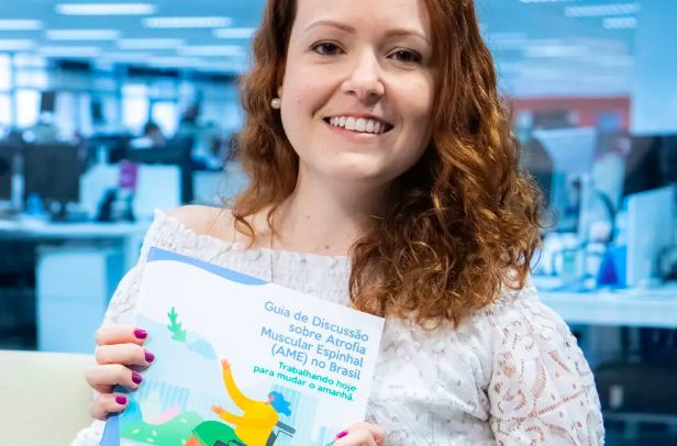 Foto de uma mulher da cintura para cima. Com ambas as mãos, ela segura um livreto intitulado “Guia de Discussão sobre a Atrofia Muscular Espinhal (AME) no Brasil: Trabalhando hoje para mudar o amanhã”. Na capa, há a ilustração de uma mulher cadeirante, com os braços erguidos e com uma planta flutuando acima dela.